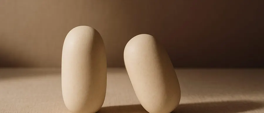 Two matching pale stone forms on warm linen, one upright and one leaning, a still life standing in for facial symmetry and imbalance